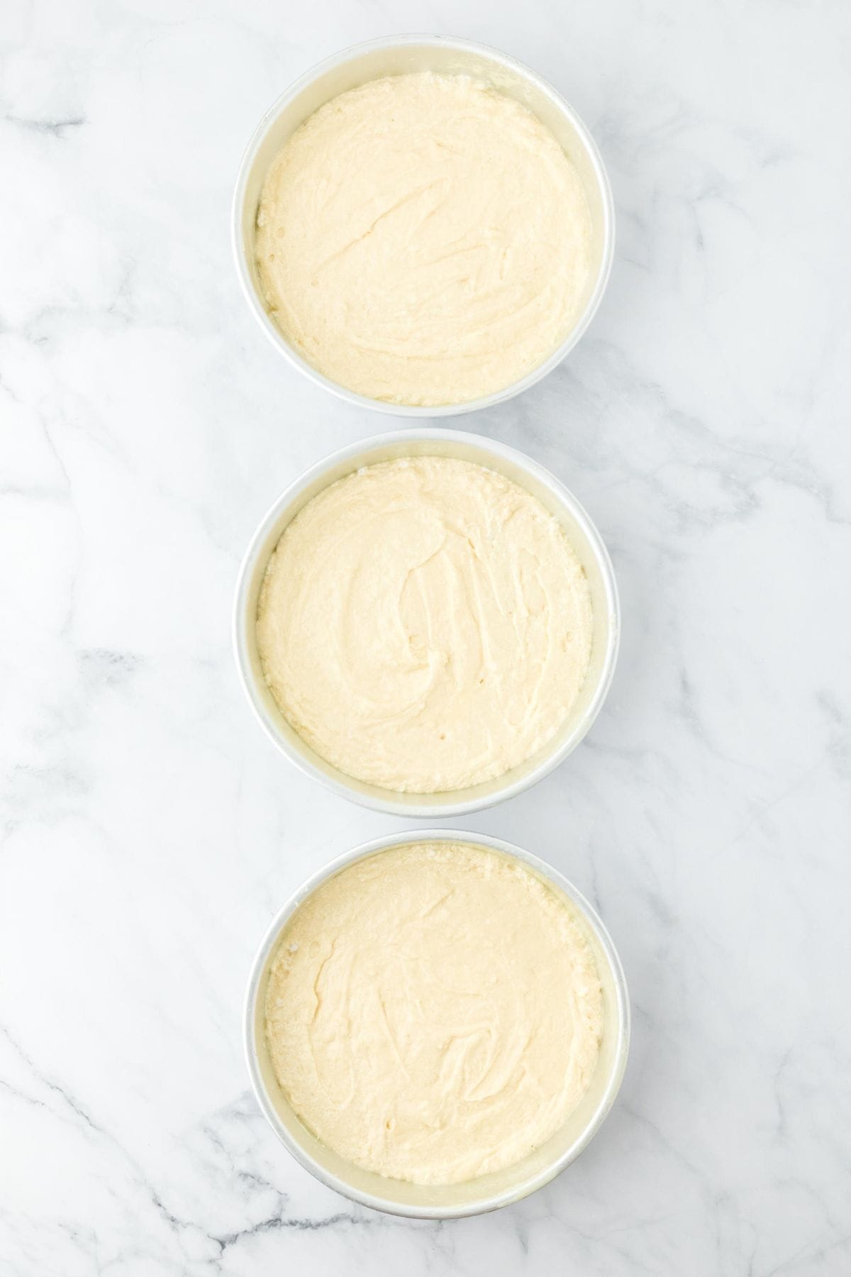 Three round pans filled with cake batter, placed on a marble surface, ready for baking