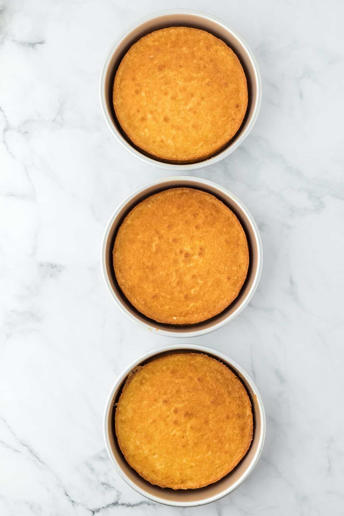 Three golden-brown cake layers in round baking pans, cooling on a marble surface