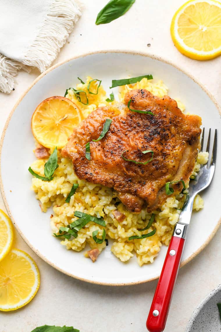 Once chicken thigh on a plate surrounded by baked lemon rice on a white plate with a red fork nearby