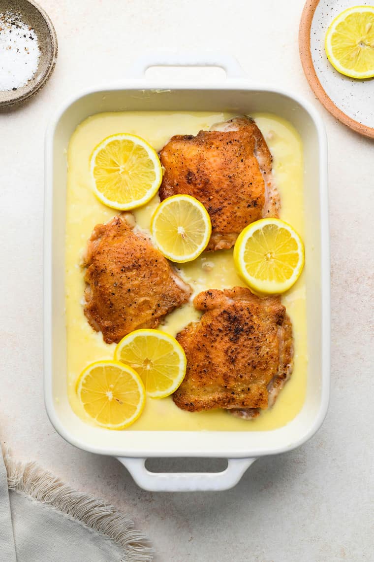 Three sauteed chicken thighs in a white casserole dish in a creamy lemon sauce with lemon slices on top