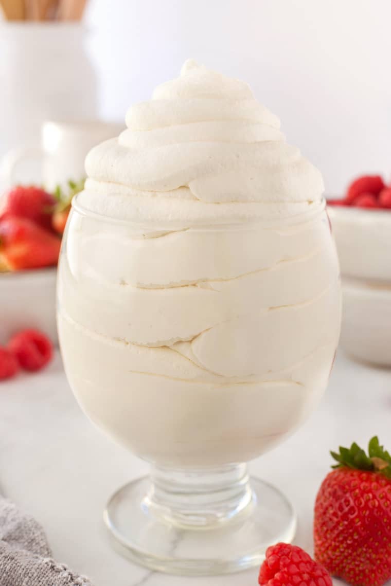 A tutorial of how to make whipped cream with whipped cream in a tall clear glass with strawberries in the background