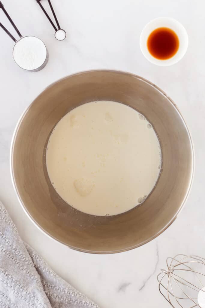Heavy whipping cream in a large silver bowl with vanilla, powdered sugar and salt nearby