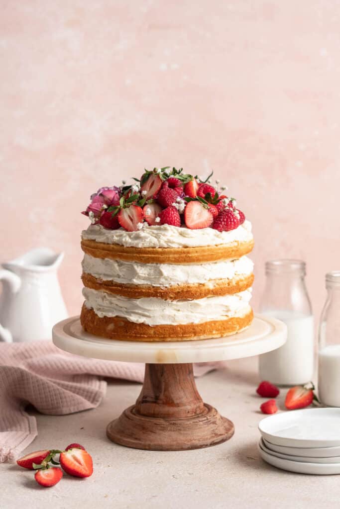 A three layer white cake on a cake stand topped with strawberries against a pink background