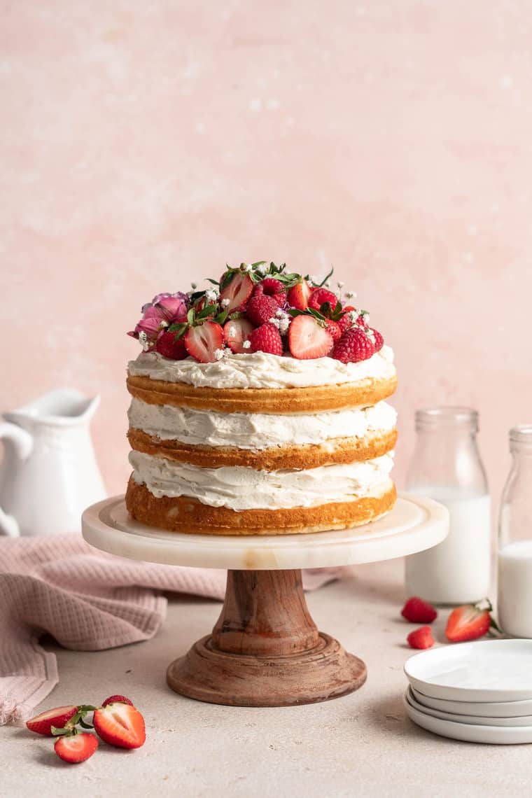 The best white cake recipe on a cake stand topped with strawberries against a pink background