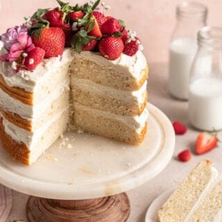 A white cake on a cake plater sliced open with a slice nearby against pink background