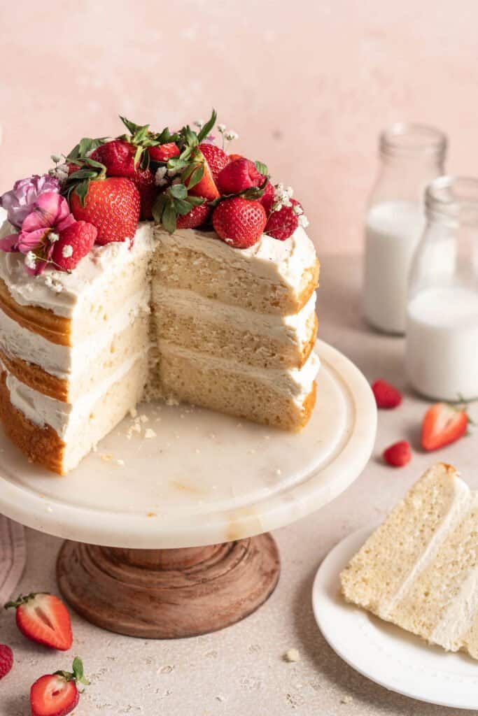 A white cake on a cake plater sliced open with a slice nearby against pink background