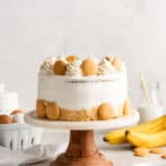 A banana pudding cake on a white and wood cake stand with nilla wafers and bananas in the background