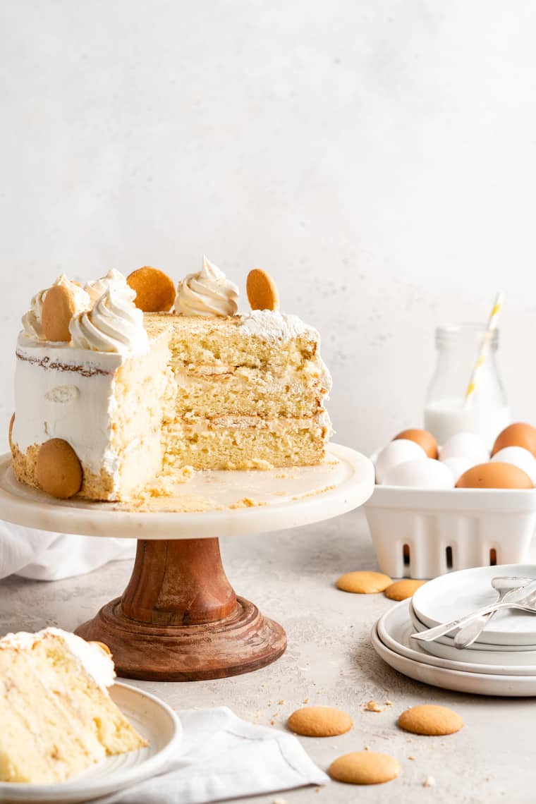 A banana pudding layer cake sliced with on a cake stand with a slice on a white plate