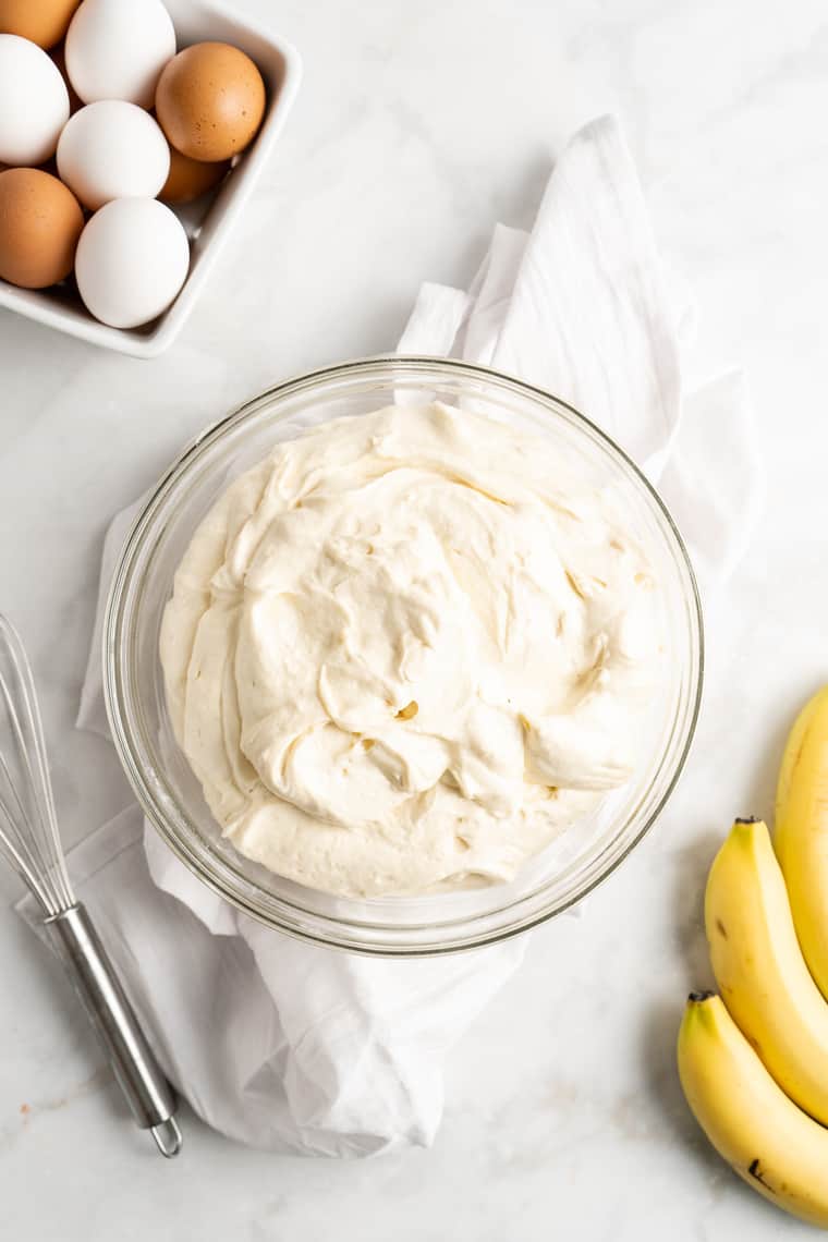 A bowl of vanilla pudding next to bananas and eggs