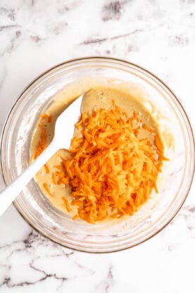 Shredded carrots being mixed into a cupcake batter with a white spatula