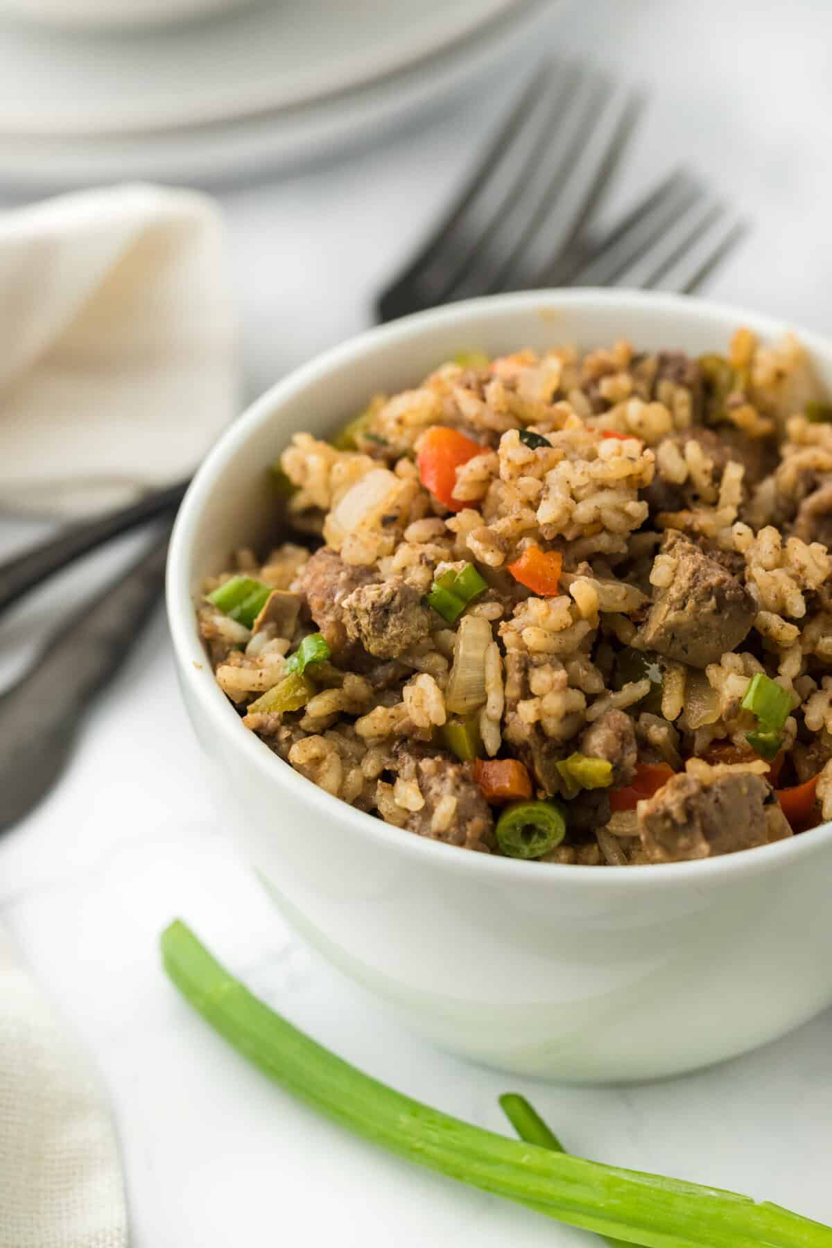 A large white bowl of Creole dirty rice on a white background with green onion and two silver forks in the background