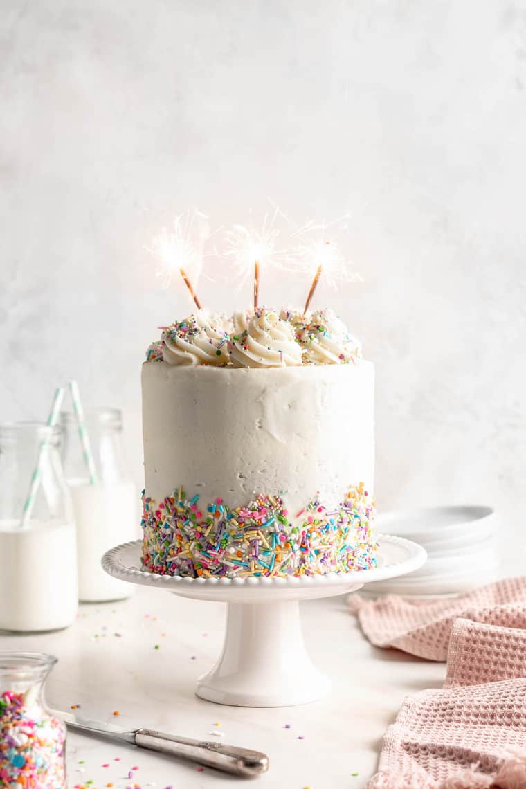 A funfetti cake with sprinkles on a white cake stand with lit candles for a birthday party