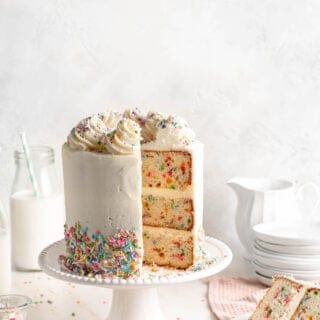 A funfetti layer cake on a white cake stand with a slice taken out on a white plate against white background