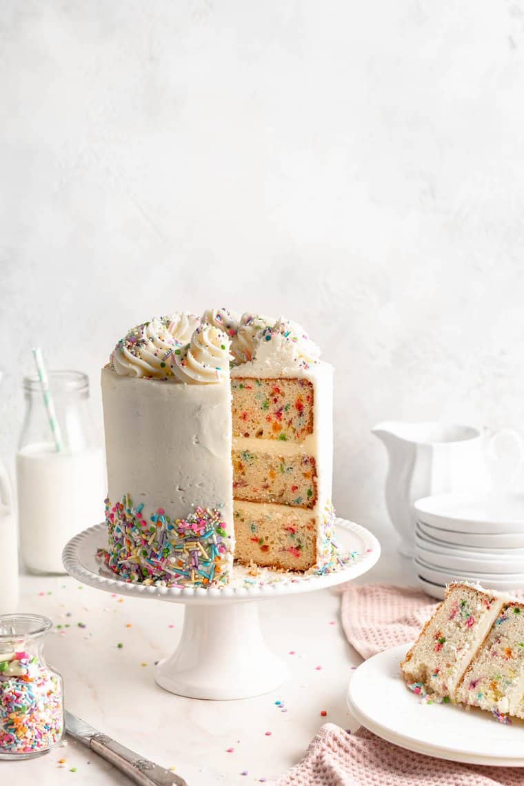 A funfetti layer cake on a white cake stand with a slice taken out on a white plate against white background