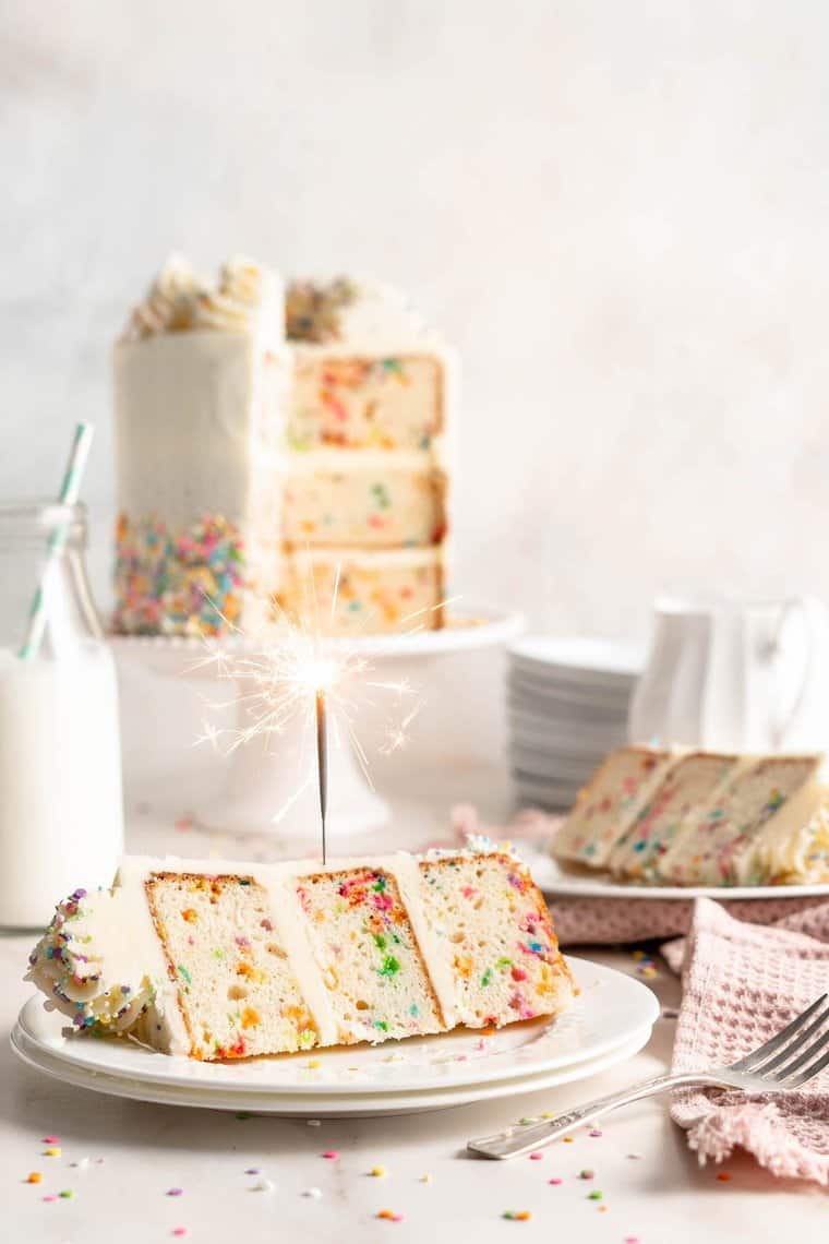 A close up of funfetti cake covered in vanilla frosting with a lit candle inserted in a slice