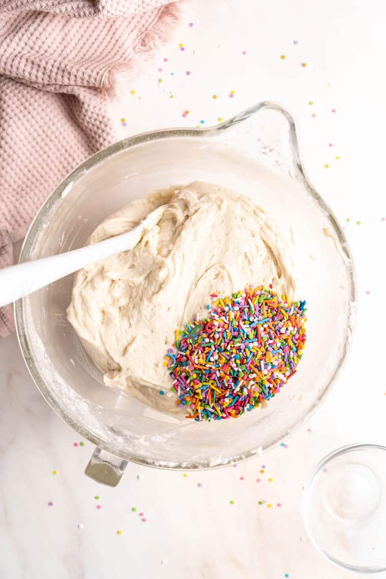 Sprinkles being stirred into a white cake batter in a clear mixing bowl