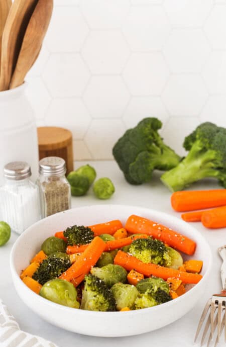 A bowl of steamed vegetables in a kitchen with chopped veggies in the background