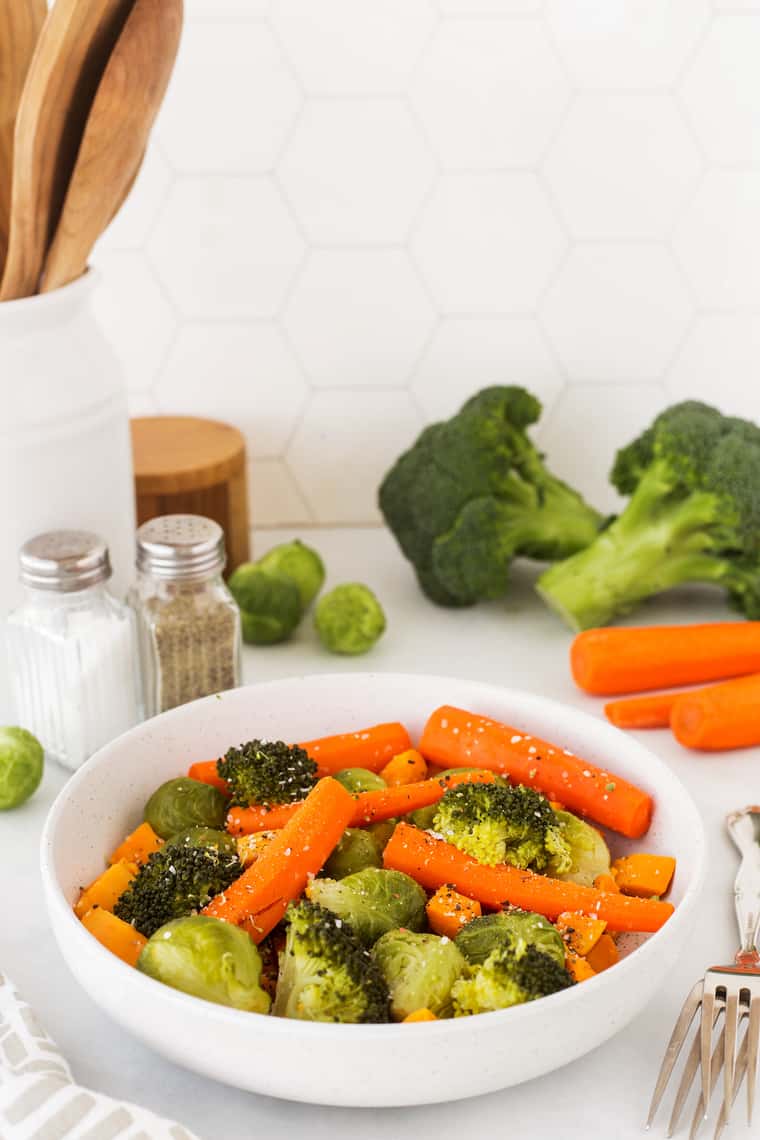 A bowl of steamed vegetables in a kitchen with chopped veggies in the background