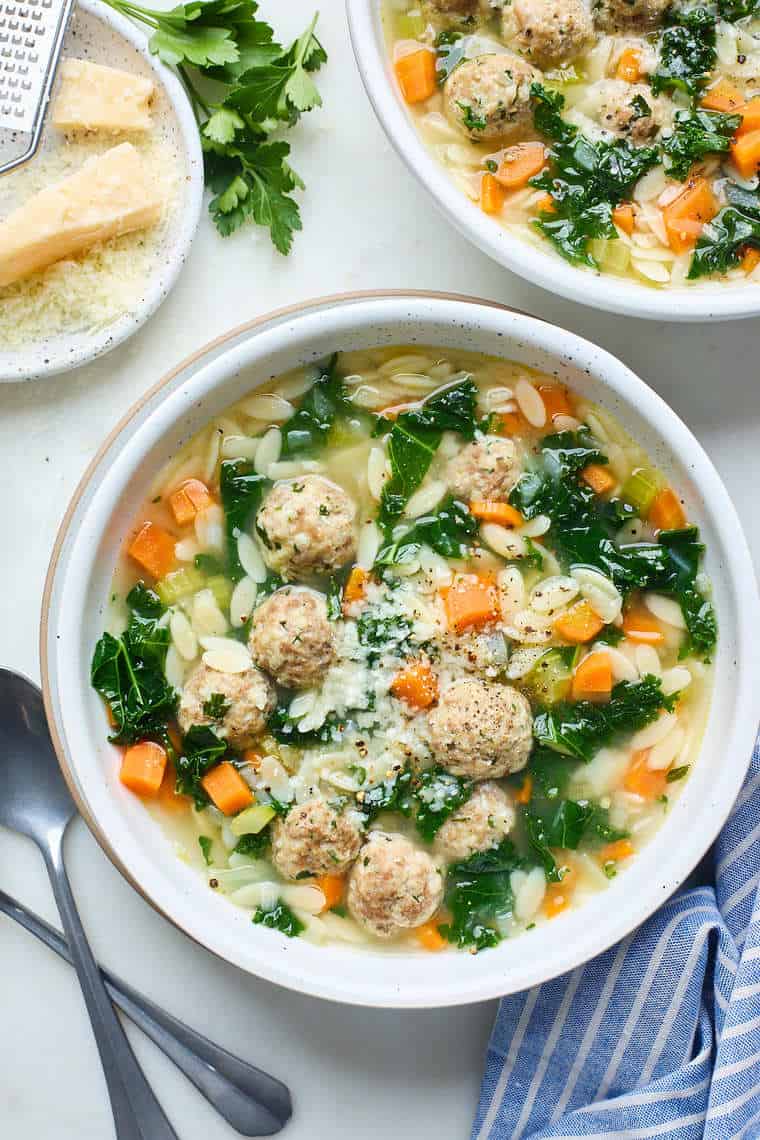 Bowls of Italian Wedding Soup against a white background with parmesan cheese in the background