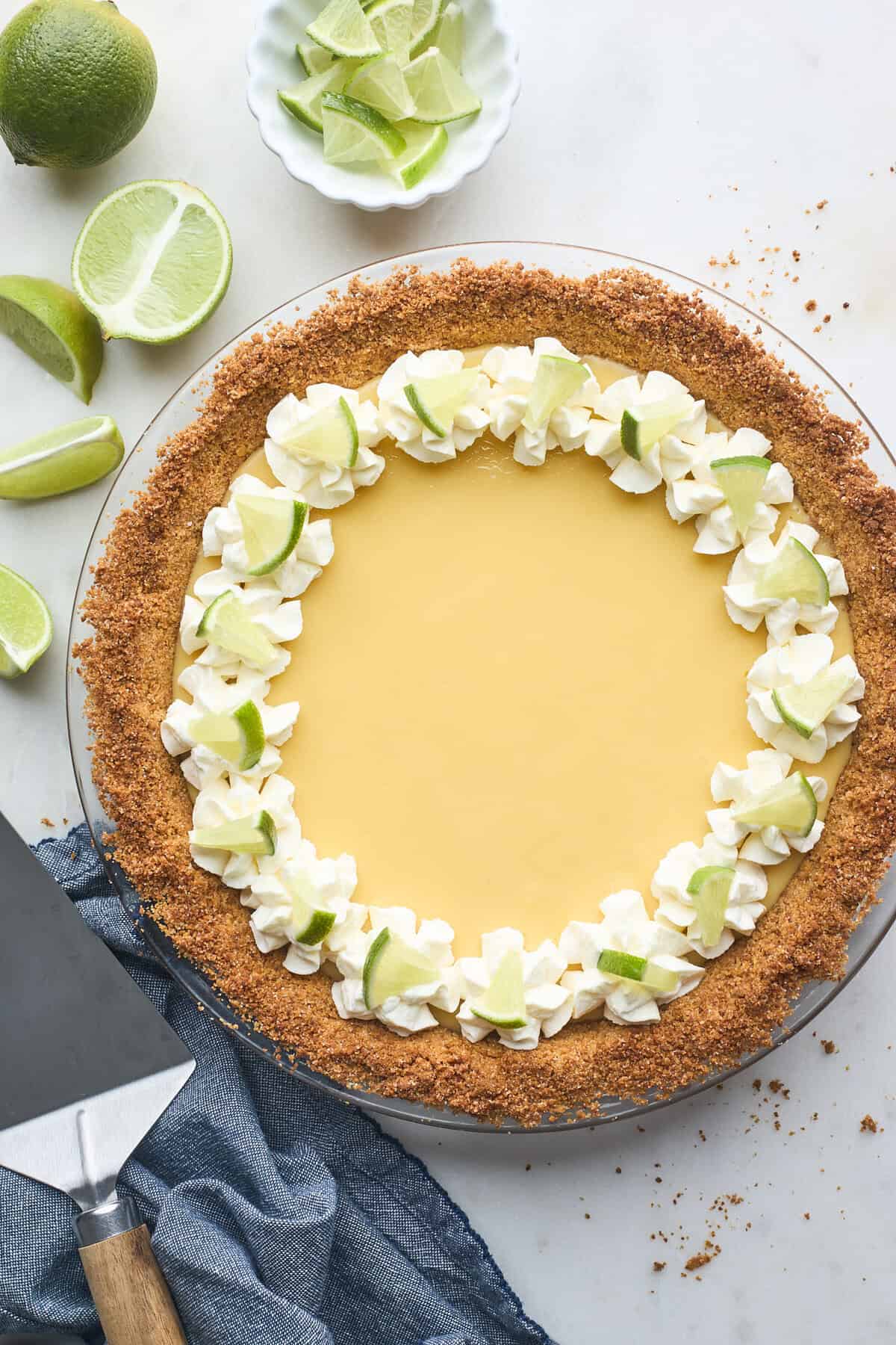 A traditional key lime pie against a gray background with key limes in the background and a spatula to serve