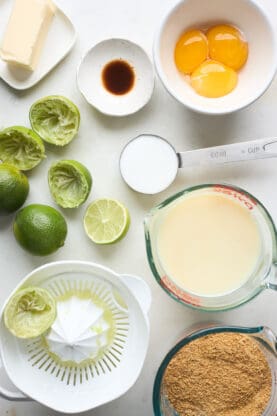 An overhead of ingredients like used key limes, egg yolks, vanilla, sweetened condensed milk and graham cracker crumbs on a white background