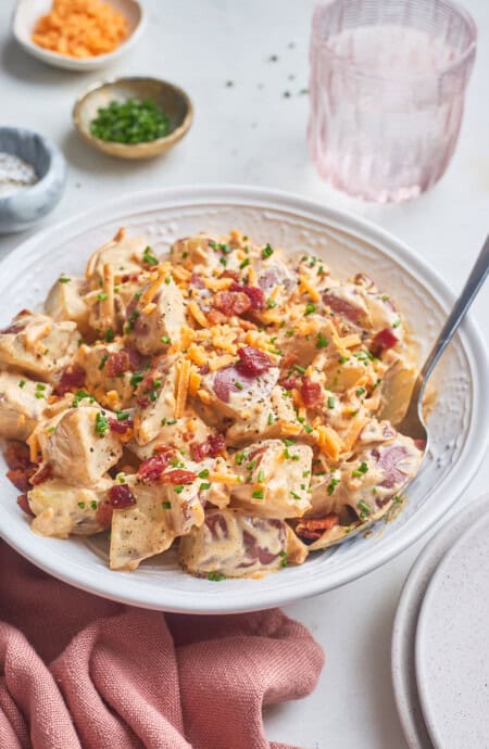 A close up of a bowl of baked potato salad with a spoon serving it