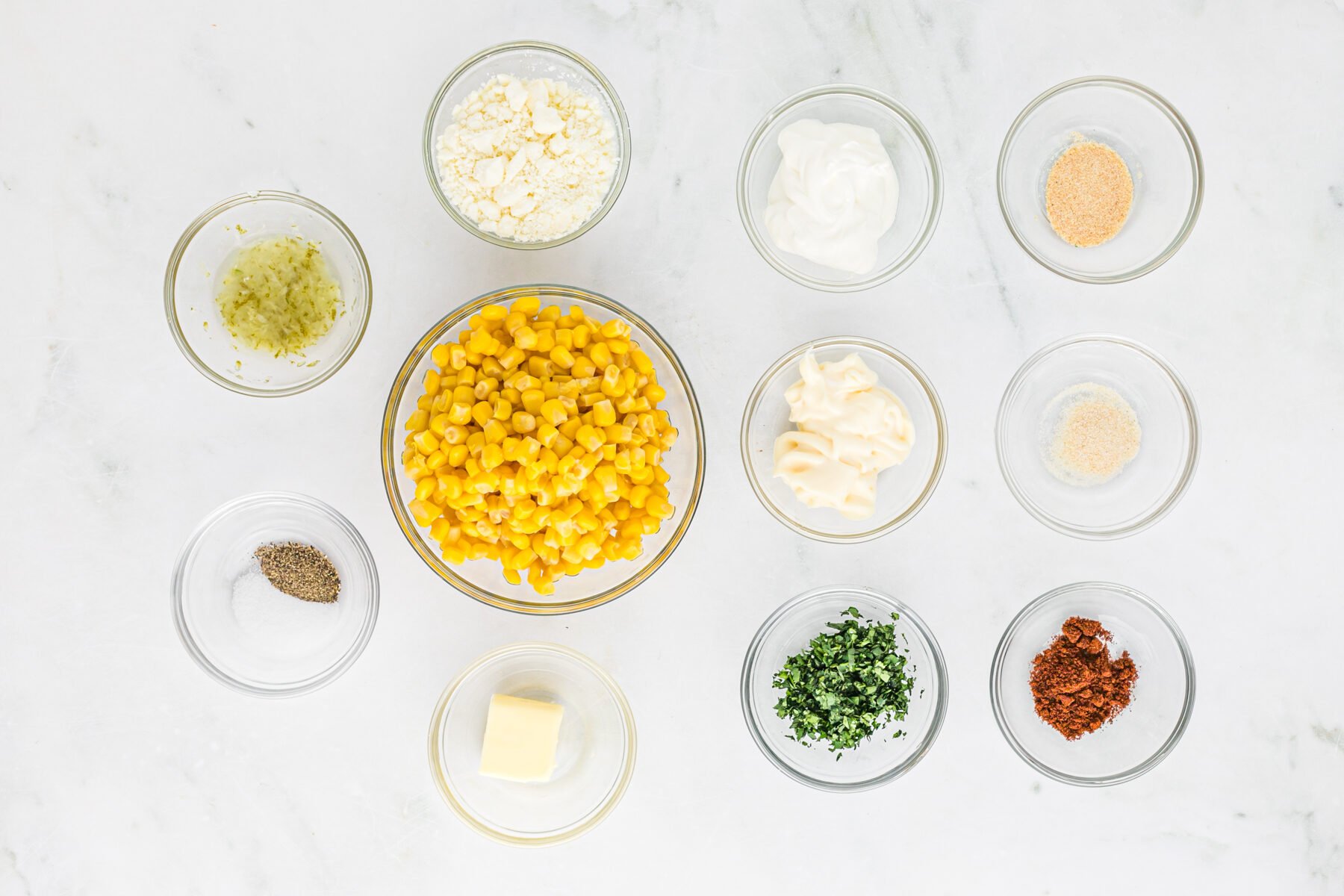 Corn, cheese, spices and cilantro in clear bowls on a white background before combining into a dip
