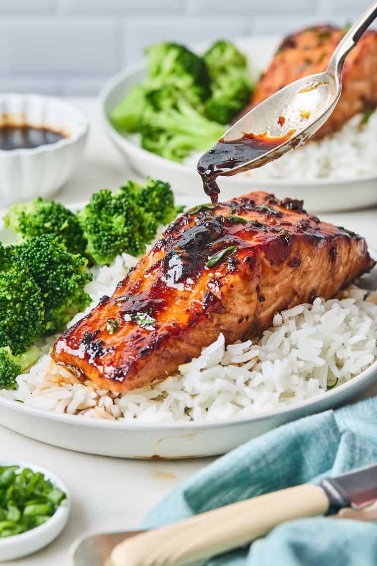 A honey garlic sauce being spooned over air fryer salmon on a plate with rice and broccoli