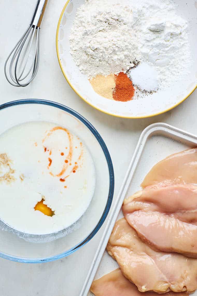 Chicken breasts, a milk mixture of hot sauce and eggs and spices and flour on a separate bowl
