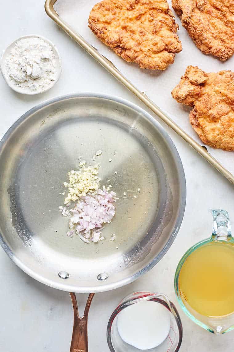 Fried chicken breasts on a baking sheet with a skillet making gravy with garlic and shallots nearby