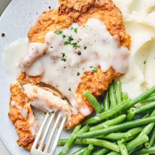 A close up of chicken fried chicken cut with a fork going into it to serve with mashed potatoes and green beans on a plate