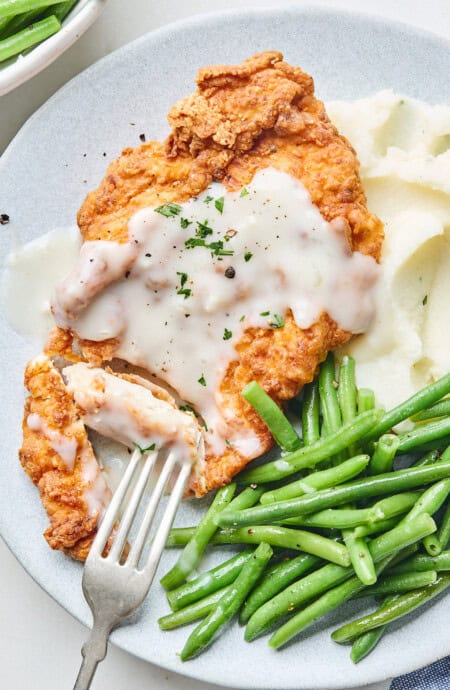 A close up of chicken fried chicken cut with a fork going into it to serve with mashed potatoes and green beans on a plate