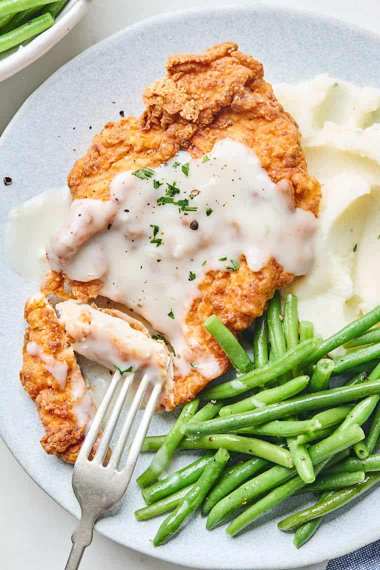 A close up of chicken fried chicken cut with a fork going into it to serve with mashed potatoes and green beans on a plate