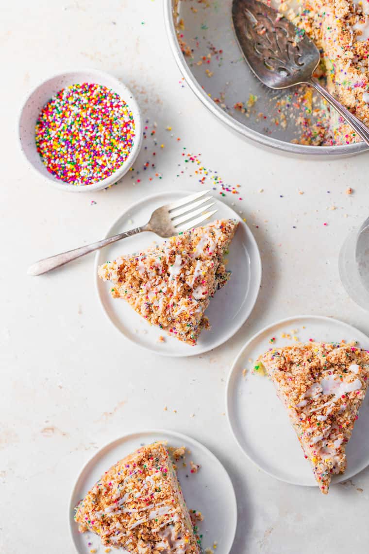 Slices of funfetti crumble cake on three plates with sprinkles in a bowl on a white surface