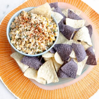 A beautiful orange plate with various tortilla chips and a bowl of elote dip ready to serve