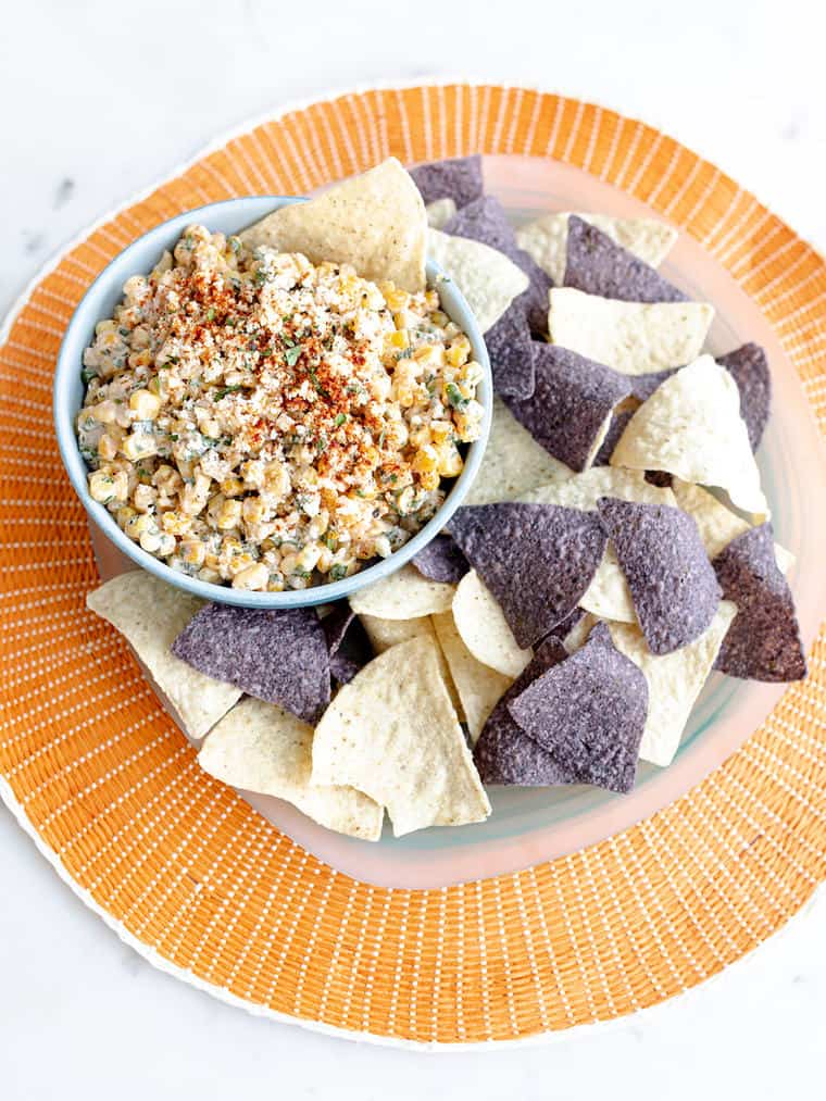 A beautiful orange plate with various tortilla chips and a bowl of elote dip ready to serve