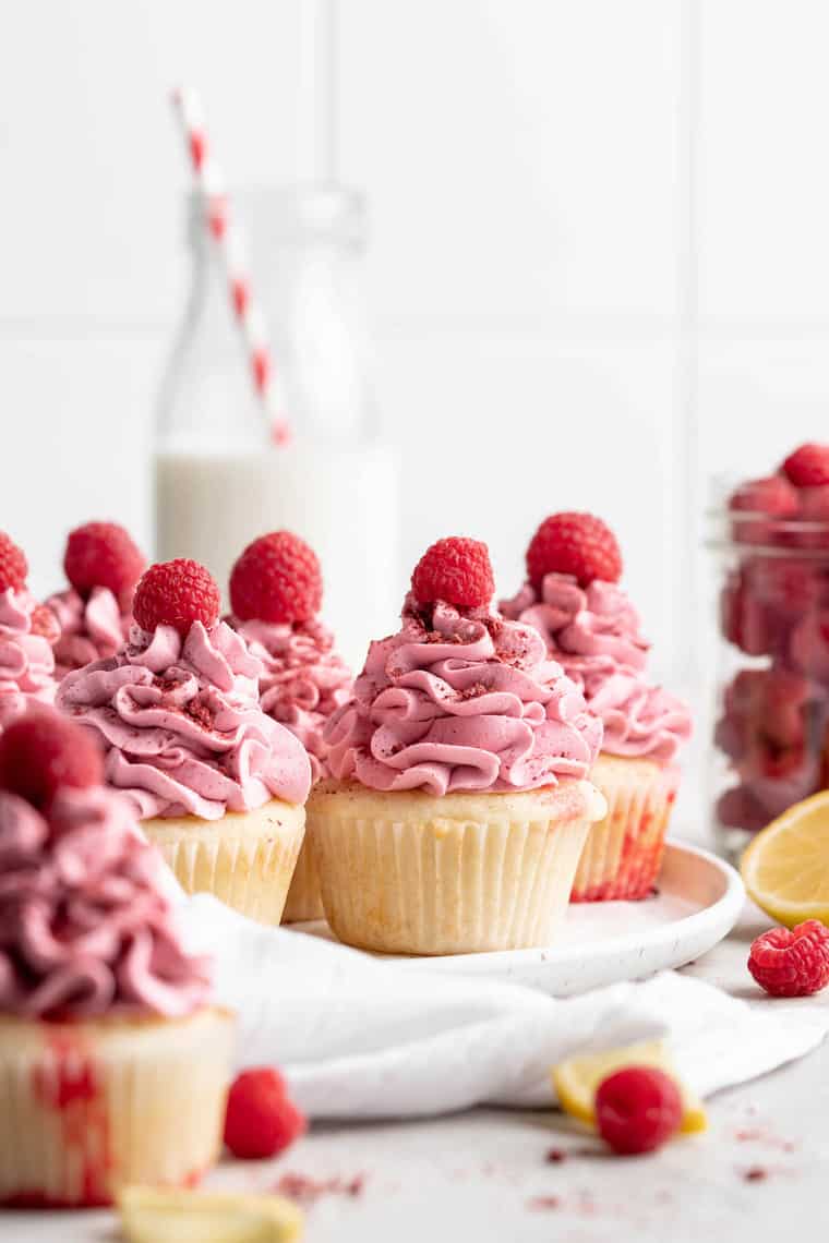 Several raspberry lemon poke cupcakes with raspberry buttercream next to each other with a glass of milk in the background