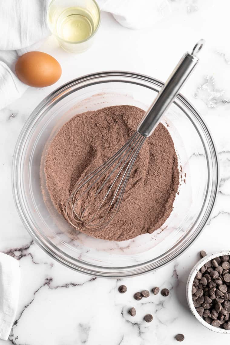 A whisk in a clear bowl with cocoa powder, flour and sugar