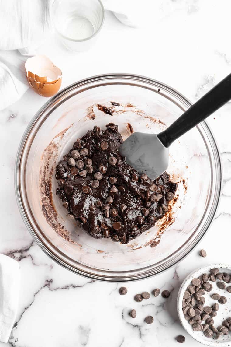 Brownie batter in a clear bowl with chocolate chips being folded in with a spatula