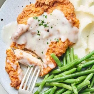 A close up of chicken fried chicken cut with a fork going into it to serve with mashed potatoes and green beans on a plate