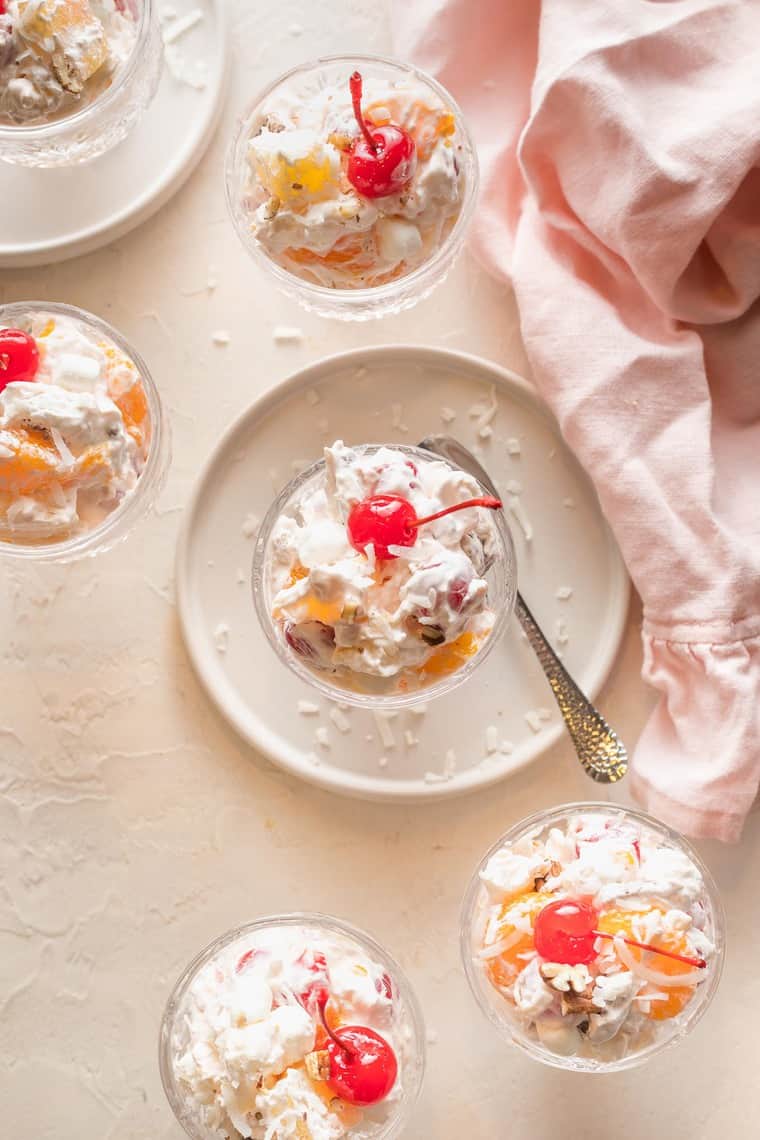 Ambrosia in small clear bowls with whipped cream and a cherry on top with a pink napkin in the background.