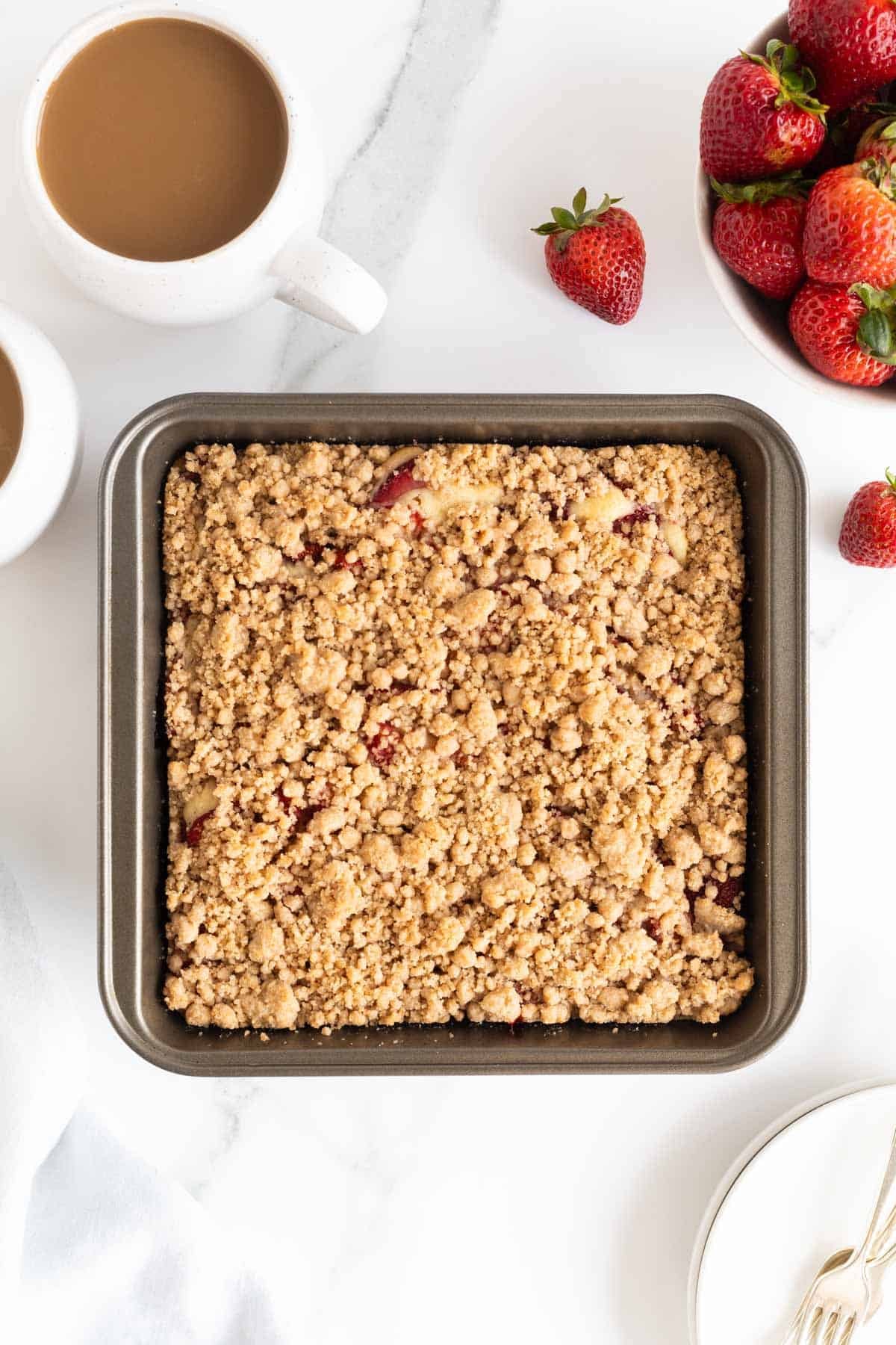 Overhead shot of a square baking pan filled with baked strawberry crumble cake, with fresh strawberries and coffee mugs around it