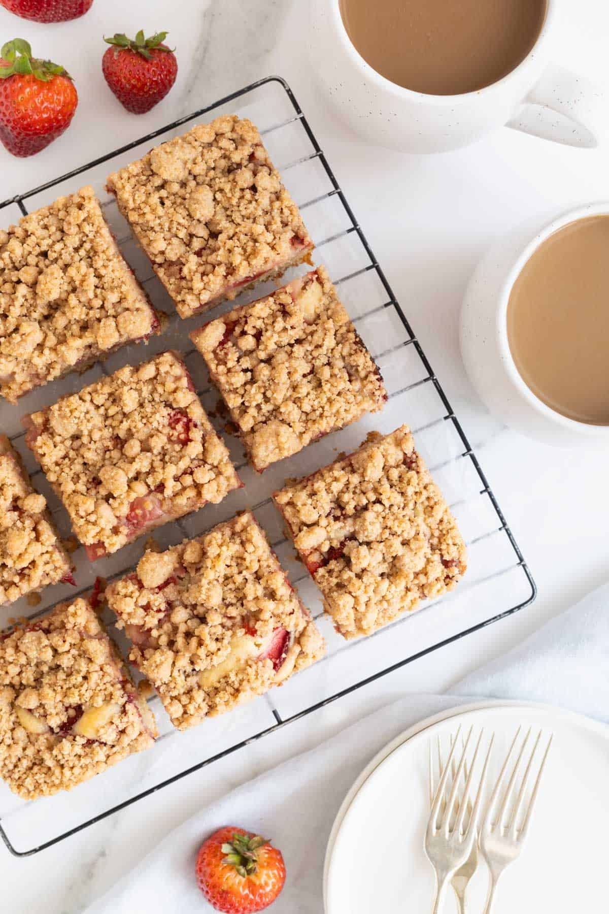 Overhead shot of several square slices of strawberry crumble cake with golden-brown topping, cooling on a black wire rack, with fresh strawberries and mugs of coffee nearby on a white marble surface