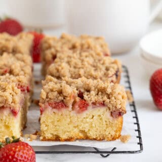Close up side view of several square slices of strawberry crumble cake on a black wire rack, with fresh strawberries in the foreground and blurred mugs in the background
