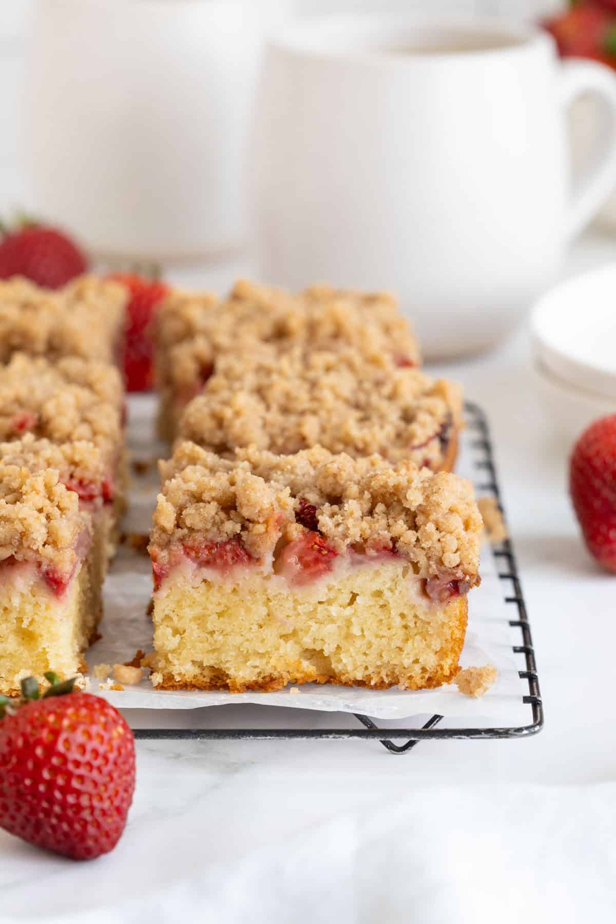 Close up side view of several square slices of strawberry crumble cake on a black wire rack, with fresh strawberries in the foreground and blurred mugs in the background