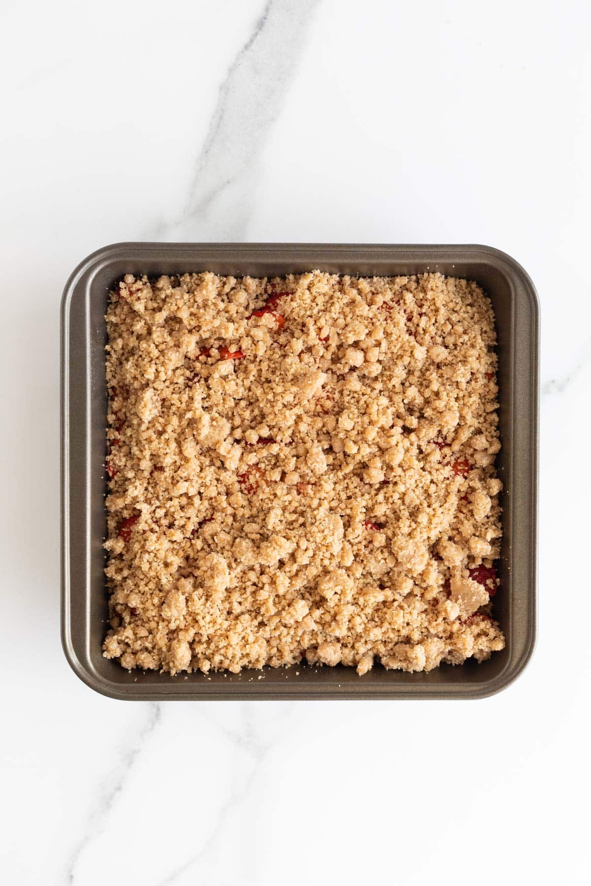 Overhead shot of a square baking pan with a layer of cake and strawberries, completely covered by a golden-brown, crumbly streusel topping, ready for baking