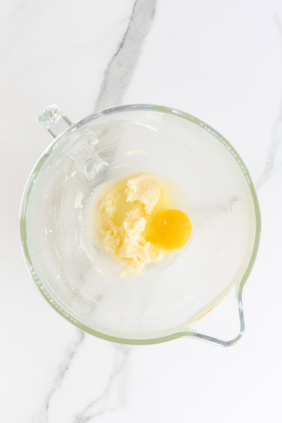 An overhead shot of a clear glass mixing bowl containing a dollop of softened butter with sugar and a raw egg yolk, on a white marble surface