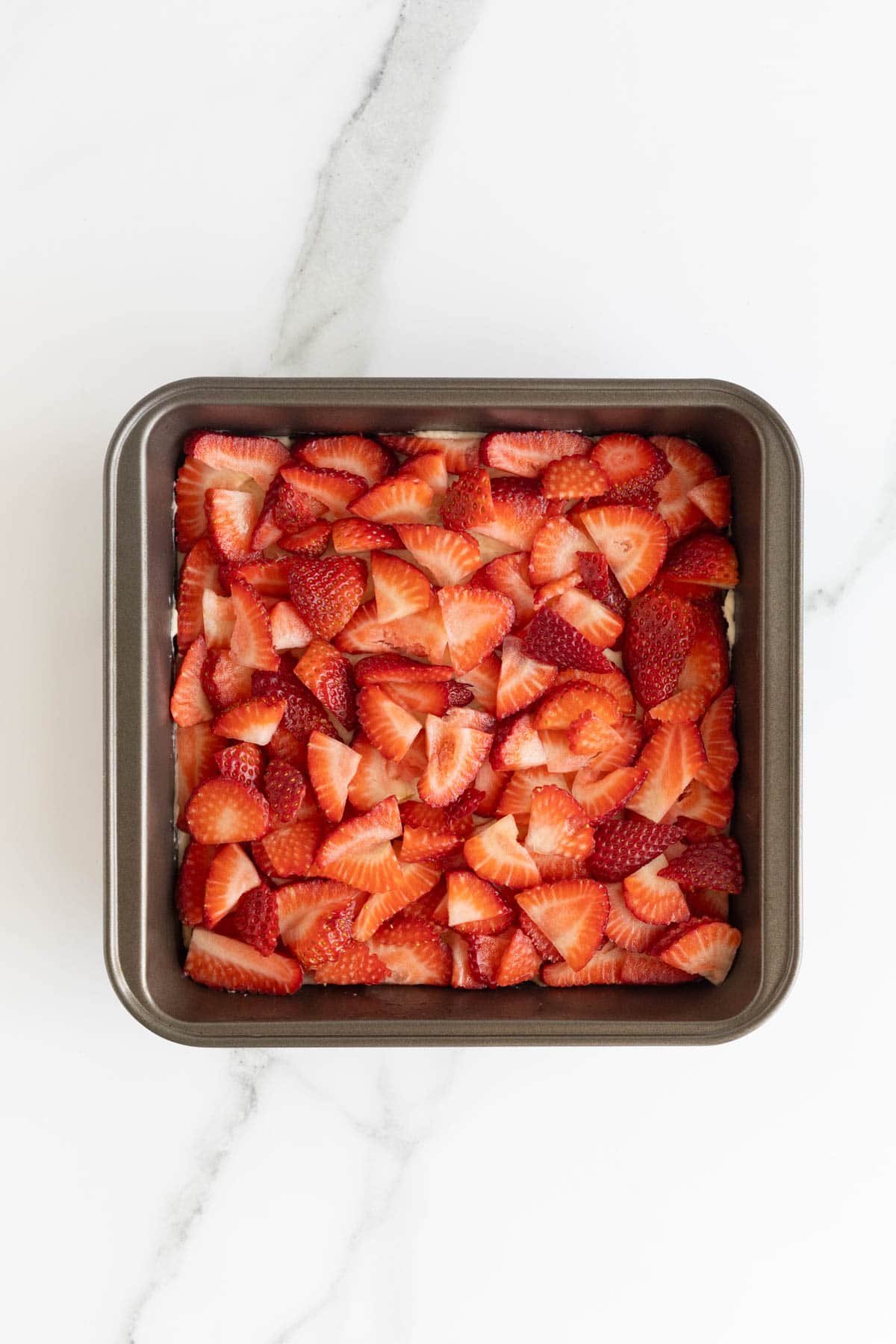 Overhead shot of a square baking pan with a layer of cake batter topped completely with a dense, even layer of chopped fresh strawberries