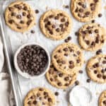 A silver baking sheet lined with parchment that has baked brown butter chocolate chip cookies on it with a bowl of chocolate chips ready to serve
