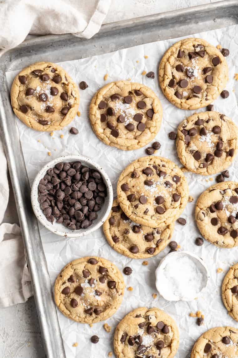 A silver baking sheet lined with parchment that has baked brown butter chocolate chip cookies on it with a bowl of chocolate chips ready to serve